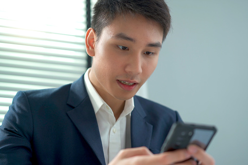 Happy cheerful Asian young businessman scrolling on smartphone screen while reading a content, happy business man using a smartphone. Man showing happy emotion after saw a good news on mobile phone.