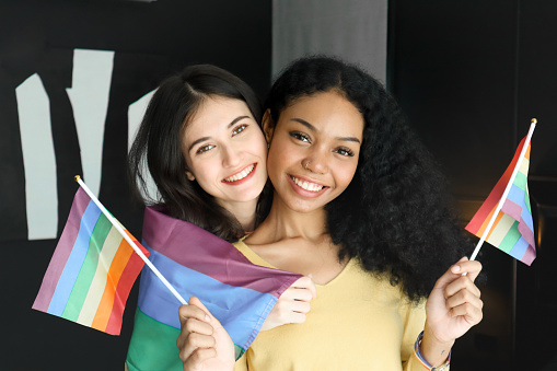 Two ethnically diverse lesbians hugging and smiling proudly about their homosexuality during pride month, attractive young women of the same gender holding rainbow flags, human rights and gender equality concepts.