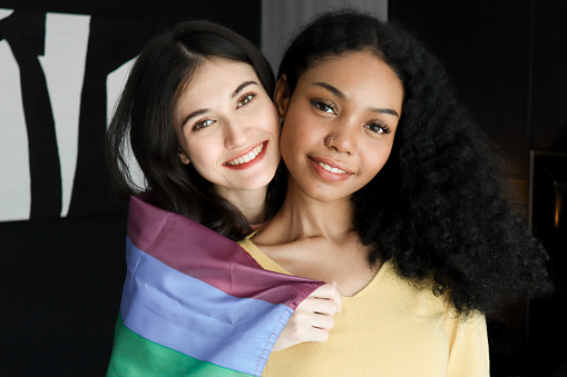 Two ethnically diverse lesbians smiling proudly about their homosexuality during pride month, attractive young women of the same gender holding rainbow flags, human rights and gender equality concepts.