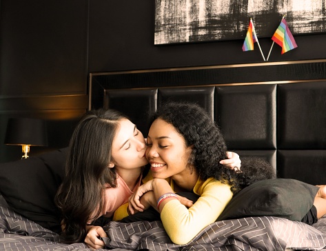 Two ethnically diverse lesbians kissing proudly about their homosexuality during pride month. Happy young women of the same gender spend romantic time in the bedroom with rainbow flags behind them.