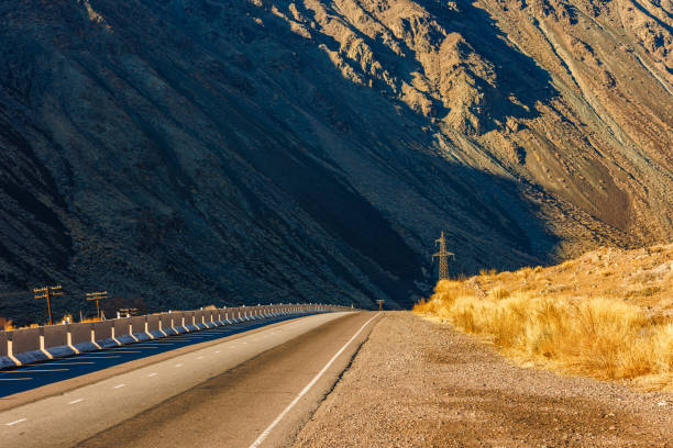 An empty highway stretches through a natural landscape with mountain in the background, showcasing the asphalt road surface as it winds through the hilly terrain.