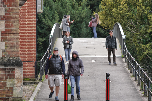 Garret Hostel Bridge, Garret Hostel Lane, Cambridge, Cambridgeshire, England – 18 March 2024: This daytime photo shows Garret Hostel Bridge crossing over Garret Hostel Lane in Cambridge. The small, believed to date back to the 15th century, connects the two halves of Garret Hostel Lane. Several people can be seen walking and standing on the bridge, enjoying the warm afternoon as part of leisure activities in the picturesque university town setting. Local people are also seen riding across the narrow bridge below trees lining the river bank. The bridge offers views up and down the tranquil river flowing through central Cambridge, a popular spot for relaxation among both locals and visitors.