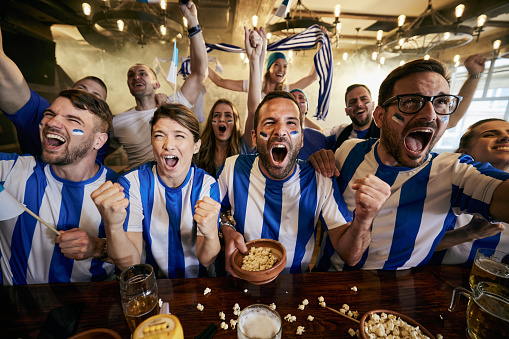 Large group of cheerful sports fans screaming while celebrating the success of their team during a game on TV in a bar.