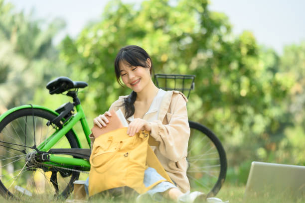 jeune femme positive emballant son sac à dos assise sur l’herbe près de son vélo dans le parc - mental health women asian ethnicity bicycle photos et images de collection