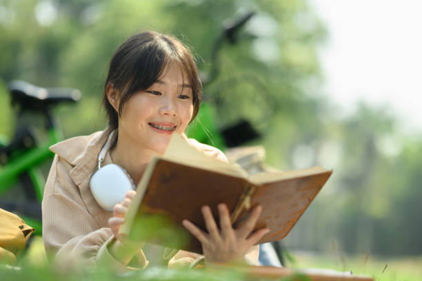 jeune femme asiatique ravie lisant un livre sur l’herbe verte pendant une journée ensoleillée - mental health women asian ethnicity bicycle photos et images de collection