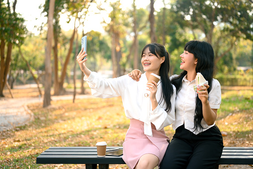 Two woman friends sitting on bench in public park and taking a selfie with mobile phone.