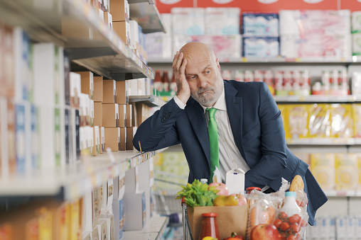 Tired confused senior businessman doing grocery shopping at the supermarket, he is leaning on the store shelves and thinking