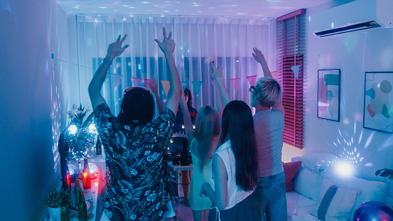 Diverse group of friends dancing moving rhythmically in good mood with EDM DJ at colorful house party at night. Multicultural friends having fun together college house party concept.