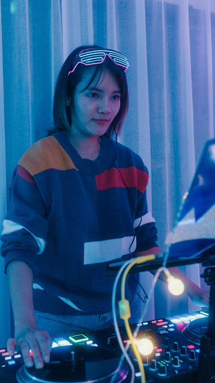 Young Asian woman dj girl playing music and scratching tracks on professional dj midi controller at house party at night. Multicultural friend having fun college house party concept. Vertical Screen.