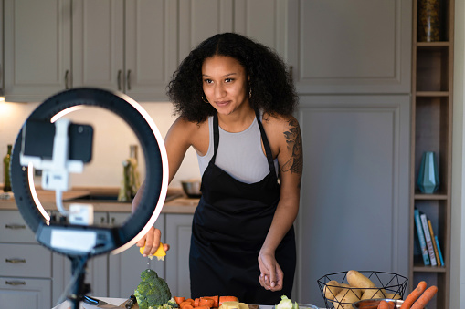 Looking at the smart phone camera held in a LED ring light, a woman is squeezing a lemon during her cooking masterclass for her online students while working from home in her domestic kitchen