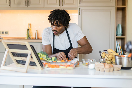 An African-American man is sitting at a table chopping fresh vegetables on a cutting board as he watches the screen on a portable device on a stand in front of him at home in his kitchen