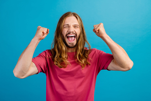 Photo of delighted ecstatic young  man raise fists shout yes success luck isolated on blue color background.