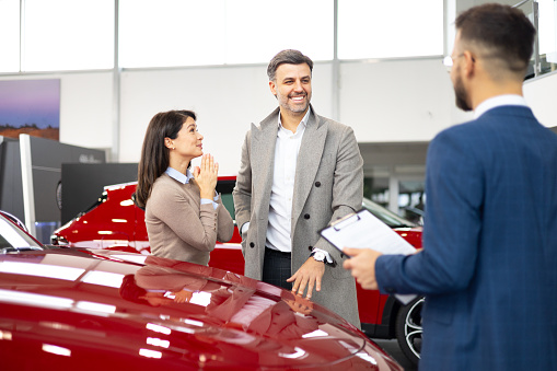 A cheerful couple engages with a car salesman in a bright car dealership, highlighting a red car