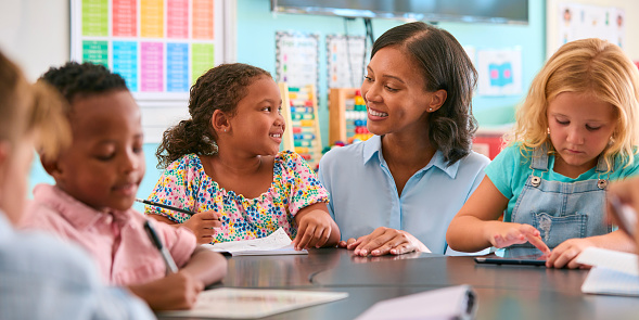 Female Primary Or Elementary School Teacher Helping Students At Desk In Multi-Cultural Class