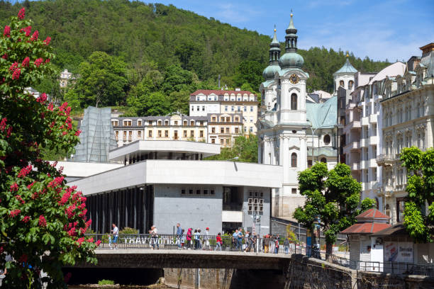 Karlovy Vary, Czech Republic -  June 3, 2023. Tourists walk on a bridge on a sunny summer day, with historical buildings and lush greenery in the background.