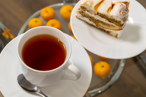 A white cup of tea sits on a table next to a slice of cake. The scene is cozy and inviting, perfect for a relaxing afternoon snack or a warm beverage