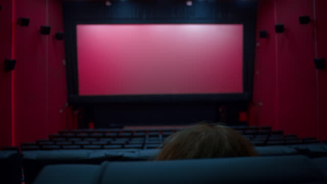 Woman taking her seat in a large empty movie theater auditorium with illumination and blank screen