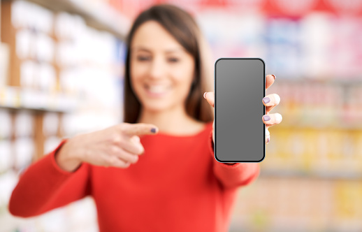 Cheerful happy woman showing her smartphone screen at the supermarket, online shopping and mobile apps concept, selective focus