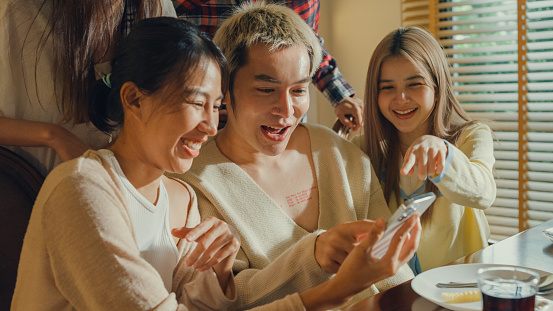 Closeup group of young Asian people watching smartphone and having fun sitting at dining table at home. Multicultural friends enjoying spending together college house party concept.