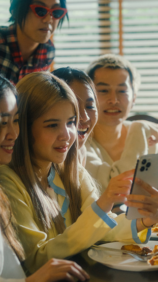Closeup group of young Asian people watching smartphone and having fun sitting at dining table at home. Multicultural friends enjoying spending together college house party concept. Vertical Screen.