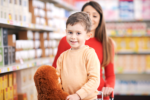 Woman pushing a shopping cart at the supermarket and cute boy standing inside, he is smiling at camera and holding a plushie