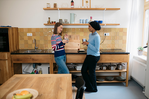 Side view of multiracial young female friends standing in the kitchen together talking and drinking coffee. Two woman roommates having coffee and talking in kitchen.