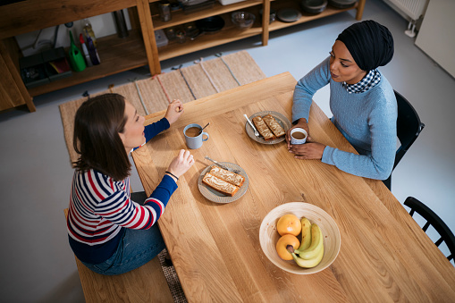 High angle view of two diverse women roommates sitting at the table in the morning having breakfast and talking. Female friends having a conversation on breakfast table in morning.