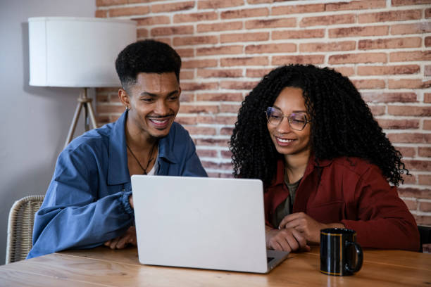 istock Handsome young couple using laptop together while sitting at home. Smiling friends looking at computer and laughing at cozy home office. 2098766858