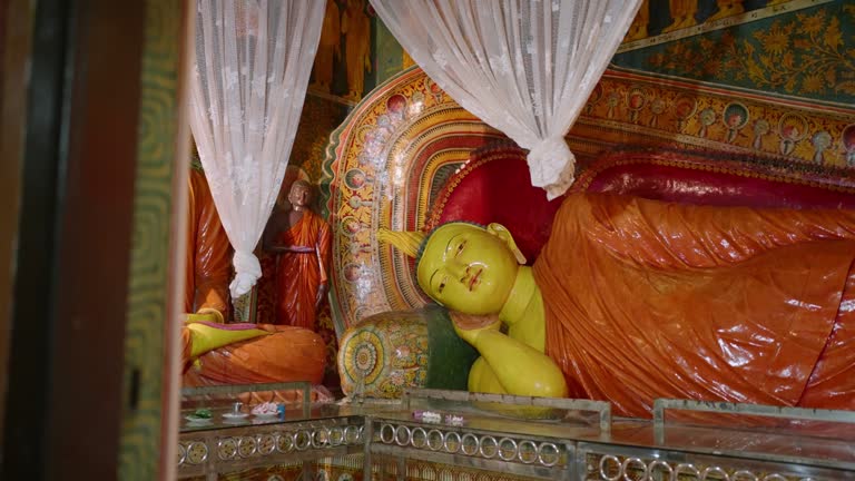 Reclining Buddha statue inside ornate temple with colorful murals. Sacred Buddhist imagery adorns walls. Tranquil spiritual setting inspires peace, meditation. Religious art reflects ancient culture.