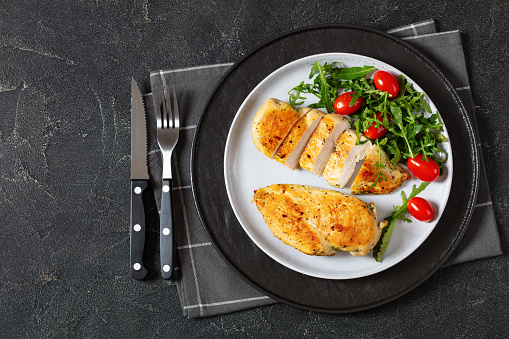 juicy fried chicken breasts with mixed green salad, tomatoes on plate on grey concrete table with cutlery, horizontal view from above, flat lay, free space