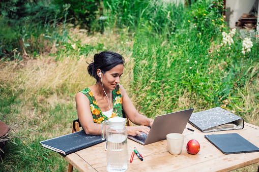 high angle view on mid adult woman sitting at wooden desk on meadow in garden and working at laptop