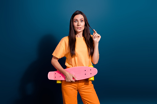 Photo of staring girl stressed biting lips touching curls brunette hair hold pink skateboard guilty isolated on dark blue color background.