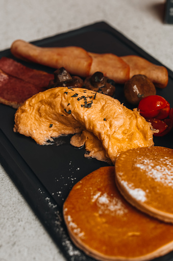 Traditional breakfast with scrambled eggs, pieces of ham, mushrooms and pancakes on a black tray