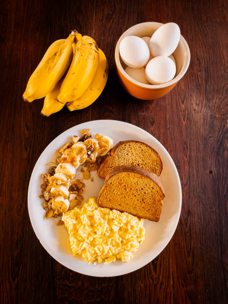 Breakfast plate set against a wooden background. The plate contains bread, sccrambled eggs, and banana with cereal and hooney, creating a wholesome and balanced morning meal.