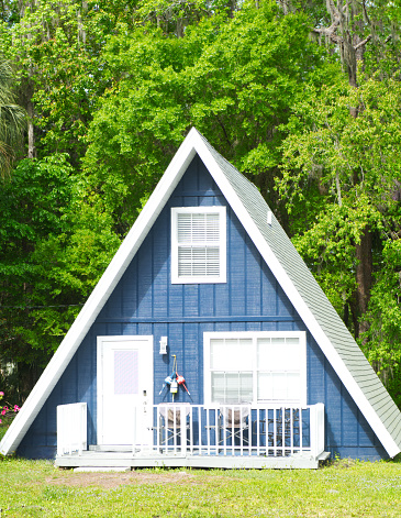 Close up of cute adorable A frame triangle shaped house or home with deep blue color, grey shingle roof, white door, wood front porch railing, nautical theme decor straight front view