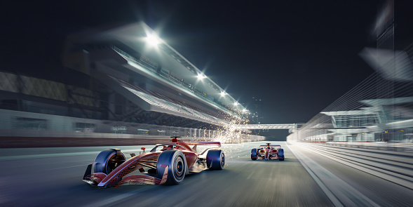 Two generic red racing cars driving at speed, one with sparks emanating from the rear along a straight past an empty spotlit grandstand at night. With motion blur to wheels and background.