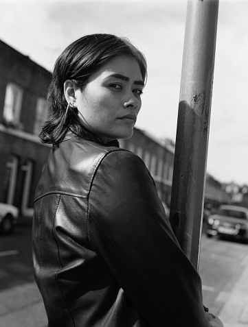 Back view of female mixed race model in a black and white portrait in the street. She is leaning against a street signal. Image made with old analog film camera.