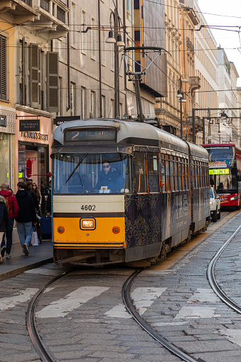 An image of a tram and its female driver who is wearing dark sunglasses. The tram is stopped at a tram stop to pick up and let off passengers. In the background you can see a red double decker bus.