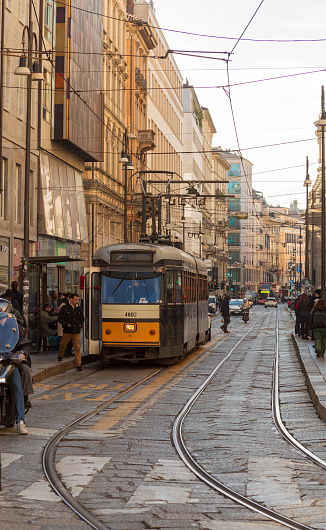 This image was taken in Milan on the 13 of Feb 2024. It shows a busy street with pedestrians and vehicles including the main point of focus, a tram which has stopped to let off and pick passengers. The image portrays normal life in the city of Milan.