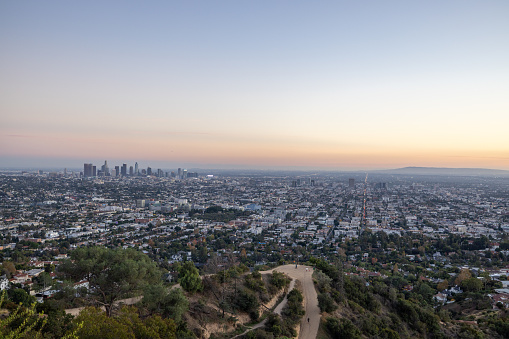 At sunset, Los Angeles transforms into a captivating vista from a high vantage point. The cityscape glows with a warm, golden hue as skyscrapers punctuate the horizon against a backdrop of vibrant oranges and pinks painted across the sky. The city's iconic landmarks stand tall amidst a sea of twinkling lights, while the fading sunlight casts long shadows that stretch across the urban sprawl below. It's a breathtaking moment where the bustling metropolis meets the serene beauty of the setting sun.