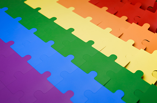 Full frame background of an LGBTQ+ pride flag shaped like a puzzle. The traditional rainbow colored pride flag where every color is its own row of puzzle pieces.