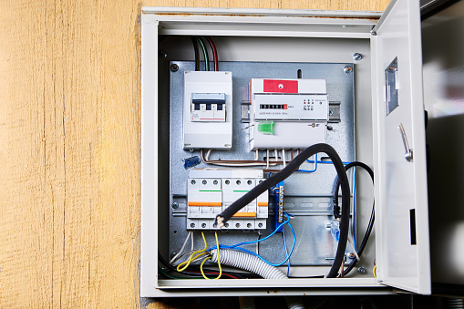 An open electrical panel is securely mounted on a wooden wall, revealing an array of circuit breakers, wires, and safety switches. The panel appears to be in the process of maintenance or inspection, with cables meticulously arranged for optimal function and safety.