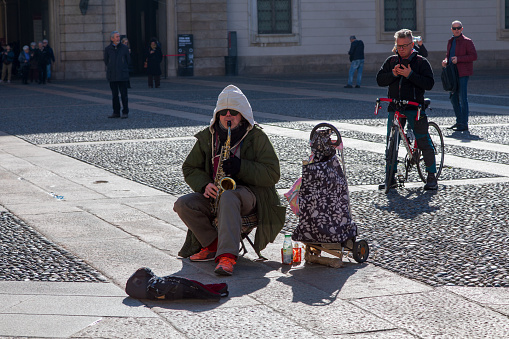 This image portrays a male street artist playing a saxophone in the Piazza del Duomo. He is wearing a green coat, a grey hoody with the hood pulled up and dark sunglasses. He is sitting on a small stool and has a shopping bag trolley next to him. Behind him is a man on a bicycle taking a photograph with two other pedestrians watching.