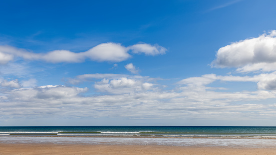 Beach of St. Andrews, Scotland, stretches out with its golden sands meeting the calm sea under a vast sky dotted with fluffy clouds, blending the hues of the ocean and the sky into a harmonious landscape