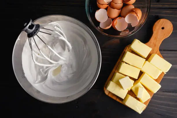 Wire whisk attachment placed in a bowl full of Swiss meringue near cracked eggs and butter
