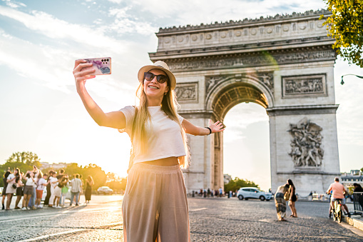 Young happy woman tourist filming vlog in front of the famous Arc de Triomphe, (Arch of Triumph) in Paris, France