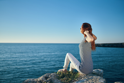 woman with hands raised up sits on high stones near the sea in summer. High quality photo