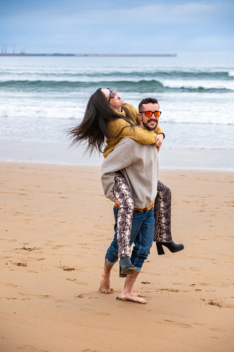 A young couple playing and laughing on the beach on a wintry day with grey skies and casual winter clothes.