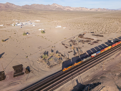 In the desert expanse of California, a freight train snakes through the arid landscape, captured from a bird's eye perspective by a drone. The train's metallic body contrasts starkly against the sandy terrain, leaving a trail of dust in its wake as it journeys across the vast desert. Against the backdrop of sun-bleached dunes and rugged mountains, the train's presence evokes a sense of both isolation and industry, a solitary traveler in a vast and unforgiving landscape.