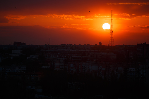 Colorful red-orange sunset with the sun's disk and the silhouette of a tower and houses in the city. Cityscape. Fiery sunset over the city.